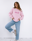A woman posing in a light pink sweatshirt with 'Pay me like a white man' printed in pink on the front, paired with light blue jeans and white sneakers, set against a plain white wall.